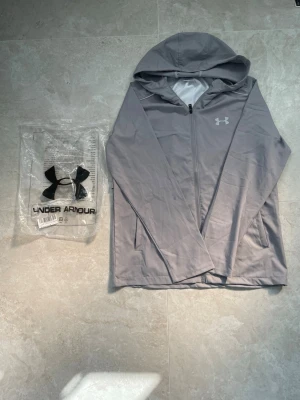 Grå vindjacka från Under Armour - Snygg grå hoodie från Under Armour med dragkedja framtill och två sidofickor. Tillverkad i ett lätt och sportigt material som passar perfekt till träning eller chill. Ikoniska UA-loggan på bröstet och huva för extra stilpoäng. Storlek:S. Helt ny med prislapp! Pris går att diskutera☺️