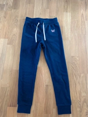 Mörkblå mjukisbyxor med fjäril - Mörkblå mjukisbyxor med vit dragsko i midjan och en liten broderad fjäril på vänster sida. Byxorna har ribbade muddar vid bensluten och är tillverkade i mjukt material för maximal komfort.