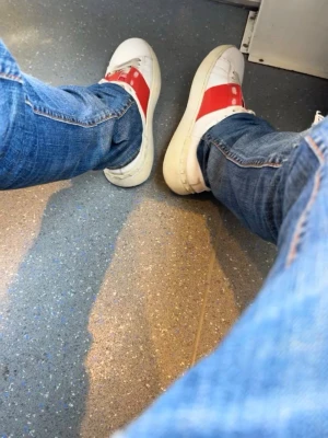 Valentino opens - Säljer ett par vita sneakers från Valentino med en bred röd remsa över mitten och nitar på hälen. Skorna har lågt skaft och är tillverkade i läder med vita snören och sula. Perfekta för dig som vill sticka ut med en ikonisk design.