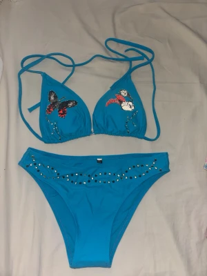 Blå bikini med pärlor och fjäril - Snygg blå bikini med knyt i sidan och dekorativa pärlor. Överdelen har broderade motiv av en fjäril och blommor i rött, svart och vitt. Underdelen är dekorerad med små pärlor framtill för extra bling. Perfekt för stranden eller poolen!