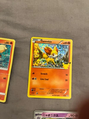 Pokémonkort Fennekin holo - Säljer ett holografiskt Pokémonkort med Fennekin. Kortet har en glittrig bakgrund och är i orange och gula toner. Det är på engelska och visar Fennekin med attackerna Scratch och Live Coal. Perfekt för samlare eller Pokémonfans.