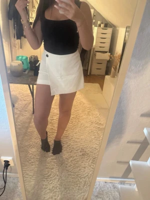 Vita shorts/kjol med knappdetalj - Säljer ett par vita shorts/kjol med snygg omlott-look och en svart knapp i midjan. Skortsen har en rak passform och är tillverkade i ett lätt tyg som passar perfekt till sommaren. Stilrena och trendiga, perfekta att matcha med crop tops eller linnen. Hög midja. Aldrig kommit till användning så i jätte bra skick.