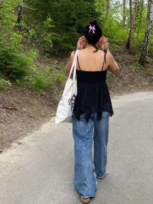 Svart omlott linnetopp med knytband - Snygg svart linnetopp från Nelly med tunna axelband som knyts på axlarna. Toppen har omlottkänsla och ett flowy, asymmetriskt fall i lätt och luftig polyester. Perfekt för varma dagar och enkel att styla med jeans eller kjol.