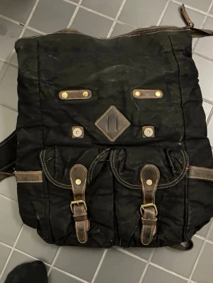 Svart ryggsäck med läderdetaljer - Rymlig svart ryggsäck med två stora framfickor och snygga bruna läderdetaljer. Justerbara axelband och dragkedja upptill. Fickor med spännen framtill och dekorativa sömmar ger en cool vintagekänsla. Perfekt för skola eller utflykter.