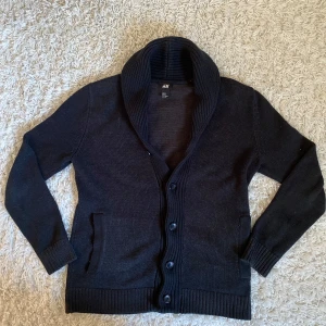 Svart stickad kofta från H&M - Snygg svart stickad kofta från H M i storlek XS. Koftan har långa ärmar, bred krage och knäpps med fyra knappar framtill. Perfekt för lager-på-lager och har två fickor framtill.