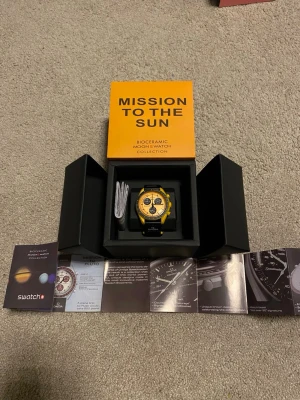 Swatch x Omega Mission To The Sun Remix - Säljer en Swatch x Omega Mission to the Sun från Bioceramic MoonSwatch Collection. Klockan har en gul boett, guldfärgad urtavla med svarta detaljer och svart armband med Omega-logga. Snygg och unik design som sticker ut på handleden.