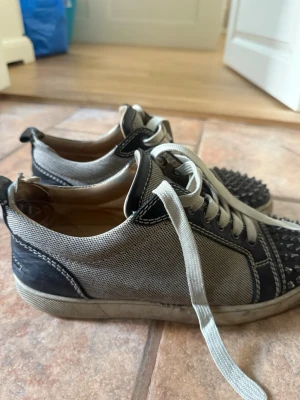 Loubs  - Säljer ett par grå sneakers från Christian Louboutin med svarta detaljer och coola nitar på tån. Skorna har vita snören och är tillverkade i canvas med läderdetaljer. Perfekta för dig som vill sticka ut med en edgy stil. Inte super bra sick därlfr priset!, inget Og 