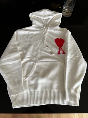 Vit hoodie från Ami Paris med rött hjärta - Säljer en vit hoodie från Ami Paris med det ikoniska röda hjärta-A trycket på bröstet. Hoodien har huva med dragsko, magficka och ribbade muddar. Materialet är mjukt och stickat, perfekt för en chill och stilren look.