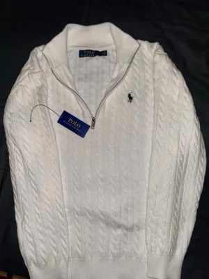 Vit kabelstickad tröja Polo Ralph Lauren - Kabelstickad vit tröja från Polo Ralph Lauren med halv dragkedja och hög krage. Tröjan har det klassiska broderade logotypen på bröstet och ribbade muddar vid ärmslut och nederkant. Perfekt för lager på lager och stilren look.