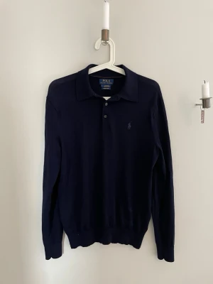 Mörkblå merinoullströja Polo Ralph Lauren - Klassisk mörkblå stickad tröja från Polo Ralph Lauren i mjuk merinoull. Tröjan har krage, knäppning med två knappar och diskret broderad logga på bröstet. Perfekt till både jeans och chinos för en stilren look.