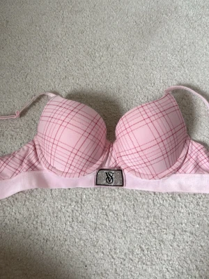 Rosa push-up bh från Victoria's Secret 75B - Söt rosa push-up bh från Victoria's Secret med rutigt mönster i rött och dekorativt VS-spänne framtill med strassdetaljer. Bh:n har vadderade kupor och justerbara axelband för bästa passform. Perfekt för dig som gillar en feminin och trendig stil.