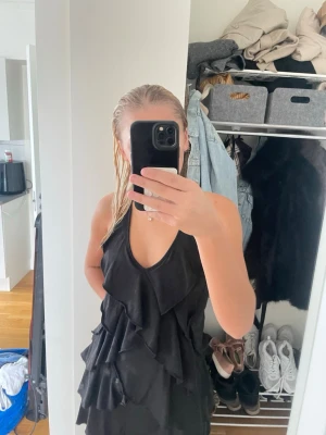 Svart volangklänning med öppen rygg - Säljer en svart kort klänning med volanger och öppen rygg. Klänningen har halterneck med knyt i nacken och ryggen, samt v-ringad front. Perfekt för dig som vill ha en stilren och trendig look.