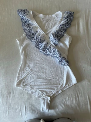 Vit body med blå spetsdetaljer från Intimissimi - Från intimissimi. Supersöt vit body med omlott-ringning och bred volangkant dekorerad med blå broderade blommor. Tunt och mjukt material, perfekt för lager på lager eller som statement-plagg. 