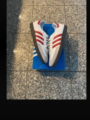 Adidas Samba vita och röda sneakers - Klassiska Adidas Samba sneakers i vitt läder med röda ränder och detaljer. Skorna har en brun gummisula och grå mocka vid tån. Ikonisk retrostil med låg profil och snörning. Perfekta för dig som gillar streetwear och sportig look. Säljer pga fel storlek.