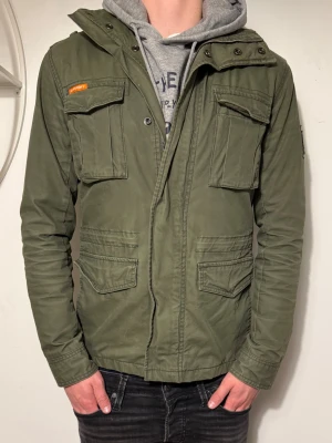 Grön vindjacka Superdry  - Snygg militärgrön vindjacka från Superdry med fyra stora fickor framtill, patchdetaljer på bröstet och ärmen. Jackan har dragkedja och tryckknappar samt camouflagemönstrat innerfoder. Perfekt för en streetwear-look. Pris kan diskuteras!💸 Strl: S✅ Hör av dig om du har funderingar!👍