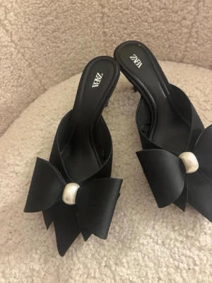 Svarta pumps med rosett från ZARA - Stilrena svarta pumps från ZARA med öppen tå och stor dekorativ rosett framtill. Skorna har en elegant pärldetalj på rosetten och en klassisk klack. Materialet ser ut att vara satinliknande tyg som ger en lyxig känsla.