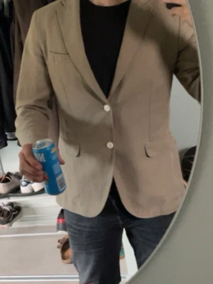 Beige kavaj med två knappar - Stilren beige kavaj med två vita knappar framtill och klassiska ficklock. Kavajen har en smal passform och enkel bröstficka. Passar M-L.