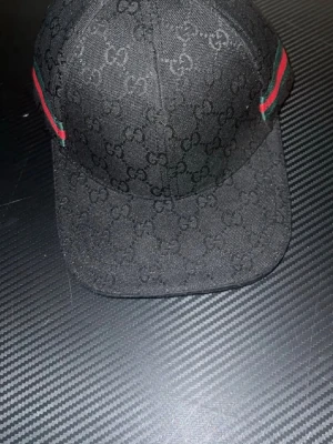 Svart keps från Gucci med GG-mönster - Snygg svart keps från Gucci med det klassiska GG-monogrammet över hela kepsen. På sidorna finns gröna och röda ränder som ger en ikonisk touch. Kepsen är tillverkad i ett slitstarkt tygmaterial och har böjd skärm.FASTPRIS! 