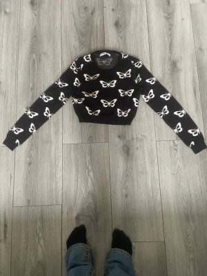Svart stickad tröja med fjärilar - Trendig svart stickad tröja med vita fjärilsmönster över hela plagget. Croppad modell med långa ärmar och ribbade muddar. Perfekt för dig som gillar unika detaljer och vill sticka ut lite extra.