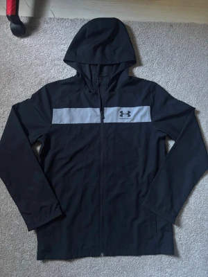 Svart vindjacka från Under Armour - Snygg svart vindjacka från Under Armour med huva och hel dragkedja framtill. Jackan har en grå panel över bröstet med logga och är tillverkad i ett lätt, vindtätt material. Perfekt för blåsiga dagar och sportiga outfits.