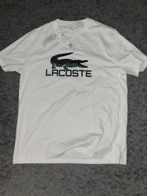 Vit Lacoste t-shirt med logga - Snygg vit t-shirt från Lacoste med den klassiska krokodilloggan och stor text på bröstet. T-shirten har rund hals och korta ärmar, perfekt för en clean och stilren look. Materialet är mjuk bomull som känns skönt mot huden.