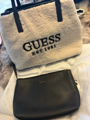 Guess handväska i teddy  - Säljer en snygg handväska från Guess i beige teddy-material med svarta detaljer och stor logga framtill. Till väskan hör en svart clutch i slätt material med guldfärgad Guess-logga och dragkedja upptill. 
