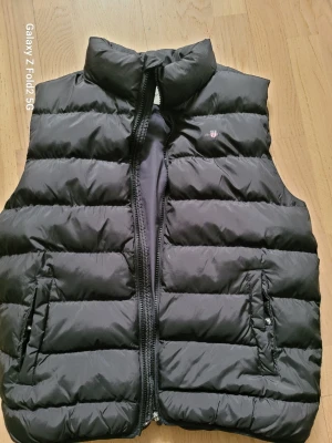 Svart dunväst från GANT - Snygg svart dunväst från GANT med hög krage och quiltad design. Västen har dragkedja framtill, två fickor med tryckknappar och GANT-logga med sköld på bröstet. Perfekt för lager-på-lager och stilren look.