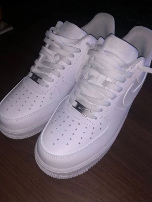 Nike Air Force 1 vita sneakers - Klassiska helvita Nike Air Force 1 sneakers i läder med perforerad tåbox, vita skosnören och diskret Nike-logga på sidan. Tidlös design med robust sula och metallbricka på snörningen där det står 'AF1'. Perfekta för en clean och stilren look.