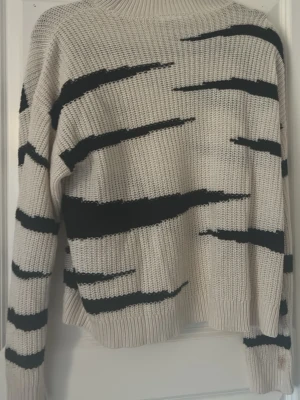 Beige stickad tröja från VILA - Säljer en beige stickad tröja från VILA med svarta, oregelbundna ränder som ger en cool och unik look. Tröjan har rund halsringning, långa ärmar och ribbade muddar. Perfekt för dig som vill ha något bekvämt men ändå trendigt.