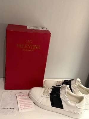 Valentino Garavani vita sneakers - Snygga vita sneakers från Valentino Garavani med svart band och nitar över vristen. Skorna är i läder och har en chunky sula samt coola detaljer baktill. Perfekta för dig som vill ha en lyxig och trendig look.