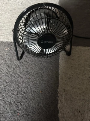 Svart bordsfläkt från Termo - Säljer en kompakt svart bordsfläkt från Termo med metallgaller och stativ. Fläkten har rund form och ansluts med USB-kabel, perfekt för skrivbordet eller mindre utrymmen. Enkel att använda med strömbrytare på baksidan.
