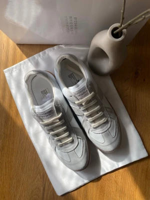 Maison Margiela vita sneakers - Stilrena vita sneakers från Maison Margiela med detaljer i grå mocka och vita läderpaneler. Klassisk låg modell med beige gummisula och vita skosnören. Perfekta för dig som gillar minimalistisk och trendig design.