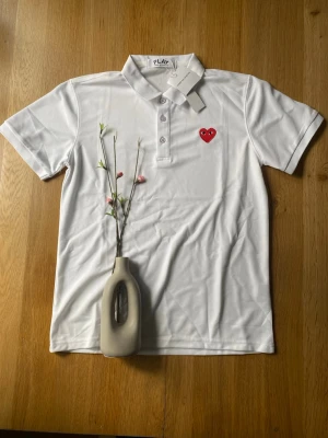 Vit pikétopp Comme des Garçons Play - Säljer en vit pikétopp från Comme des Garçons Play med klassisk krage och knappar framtill. På bröstet sitter det ikoniska röda hjärtat med ögon. Toppen är i bomull och har korta ärmar. Perfekt för en clean och stilren look.