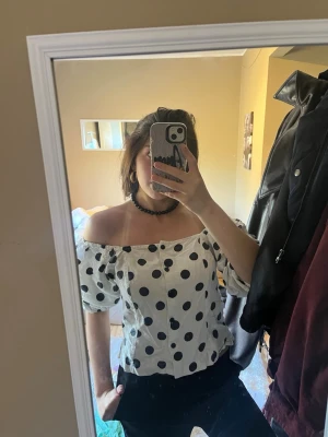 Vit offshoulder prickig blus - Säljer en vit offshoulder blus med stora svarta prickar och puffiga korta ärmar. Blusen har en feminin och trendig look med smockad resår upptill och lätt figurnära passform. 