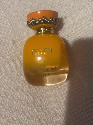 Aura Mango Splash parfym - Aura Mango Splash från La Fede är en parfym i en rund, orange glasflaska med guldfärgad dekor och orange kork. Flaskan har en lyxig och färgstark design som verkligen sticker ut. Perfekt för dig som gillar fruktiga dofter och snygga flaskor.