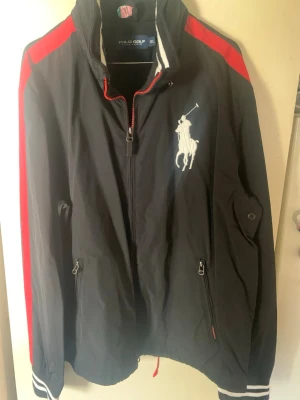 Svart vindjacka Polo Golf XL - Svart vindjacka från Polo Golf Ralph Lauren i storlek XL. Jackan har röda detaljer på axlarna, vit broderad logga på bröstet och dragkedja framtill. Ribbstickade muddar med vita ränder och två fickor med dragkedja. Perfekt för en sportig look.