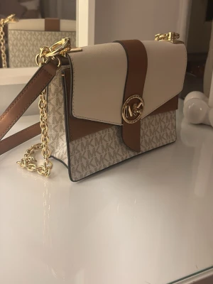 Michael Kors beige och brun handväska - Snygg handväska från Michael Kors i beige och brunt med MK-logga framtill. Väskan har guldfärgade detaljer, kedjerem och klassiskt MK-mönster på nedre delen. Tillverkad i konstläder och canvas, perfekt för dig som gillar stilrena accessoarer.