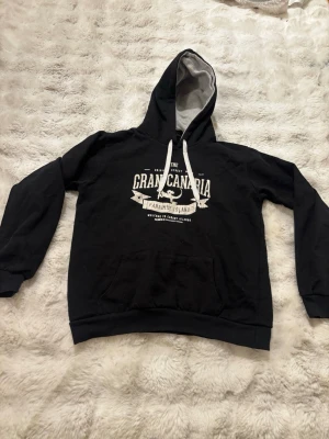 Svart hoodie Gran Canaria print - Svart hoodie med vit text och tryck 'Gran Canaria Paradise Island' på bröstet. Mjuk insida, vit dragsko och fodrad huva i grått. Stor känguruficka framtill. Perfekt för chill dagar eller när du vill ha en avslappnad stil.