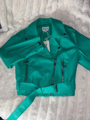 Turkos skinnjacka från NA-KD - Snygg turkos skinnjacka från NA-KD med klassisk bikerstil, silvriga dragkedjor och bälte i midjan. Jackan har bred krage, axelklaffar och två framfickor med dragkedja. Perfekt statement-plagg för dig som vill sticka ut.