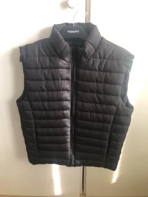 Väst  - Snygg svart dunväst från KappAhl med quiltad design och hög krage. Västen har dragkedja framtill och är lätt vadderad för extra värme. Perfekt att slänga över en hoodie eller tröja för en chill och stilren look.