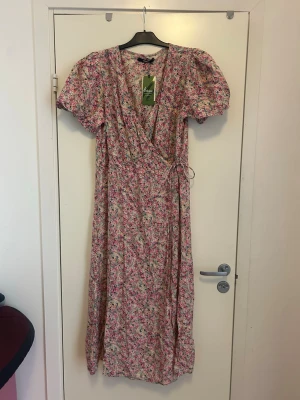 Blommig omlottklänning från Bik Bok - Söt långklänning från Bik bok med blommigt mönster i rosa, grönt och blått. Klänningen har omlottmodell med knyt i midjan, puffiga kortärmar och v-ringning. Perfekt för sommarens alla tillfällen.