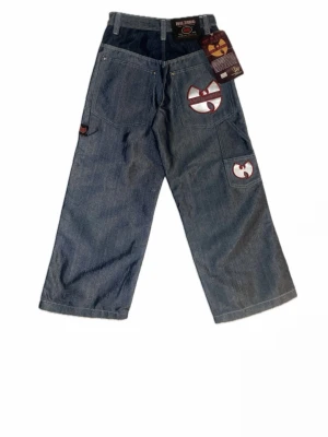 Wu-Tang baggy jeans med brodyr - Baggy jeans i blå denim från Wu-Tang med bred passform och stora fickor. Snygga broderade Wu-Tang-loggor i vitt och rött på bakfickan och benet. Klassisk femficksmodell med hällor för bälte. Perfekta för dig som gillar streetwear och unik stil.