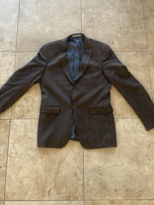 Mörkgrå klassisk kavaj herr - Stilren mörkgrå kavaj med två knappar och klassisk passform. Kavajen har två fickor med lock framtill och en bröstficka. Fodrad insida i blått tyg och enkel slits bak. Perfekt för en dressad look.