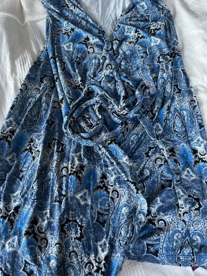 Blå omlottklänning med paisleymönster - Säljer en blå omlottklänning med paisleymönster i olika blå och vita nyanser. Klänningen har knytband i midjan och v-ringad hals. Tyget känns mjukt och följsamt, perfekt för sommarens varma dagar. Storlek  M🥰