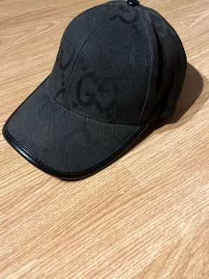 Svart keps från Gucci med GG-mönster - Snygg svart keps från Gucci med det klassiska GG-mönstret ton-i-ton över hela kepsen. Kepsen har böjd skärm med svart kant i läderimitation och justerbar rem bak. Tillverkad i ett tåligt tyg som ger en exklusiv känsla.