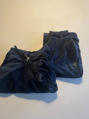 Mörkblå velourset från Juicy Couture - Säljer ett ikoniskt mörkblått velourset från Juicy Couture med glittrande Juicy-text i strass på både tröjan och byxorna. Setet har dragkedja, huva och muddar på tröjan samt dragsko i midjan på byxorna. Perfekt för en avslappnad och trendig look.