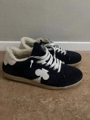 Mörkblå sneakers med fuskpäls - Snygga mörkblå sneakers med vit sula, vita skosnören och vit detalj i form av en klöver på sidan. Insidan är fodrad med mjuk fuskpäls för extra komfort. Yttermaterialet ser ut att vara mocka eller liknande tyg. Perfekta för dig som vill ha något unikt.