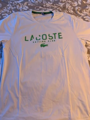Vit Lacoste Sailing Club t-shirt - Snygg vit t-shirt från Lacoste med grönt tryck på bröstet där det står 'Lacoste Sailing Club' och den klassiska krokodilloggan. T-shirten har rund hals och korta ärmar, perfekt för en clean och sportig look.