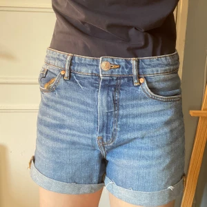 Blå denimshorts från Kappahl - Snygga blå denimshorts från Kappahl med klassisk femficksdesign och uppvikta benslut. Shortsen har hög midja och stängs med knapp och dragkedja. Perfekta för varma dagar och enkla att matcha med olika toppar. Midjan sitter högt upp och kan ge en 80-tals look om man matchar den rätt. Ifall man viker ner kanterna så kan man klä ut sig till Will Byers från Stranger Things i säsong 3 om man vill 💡. 