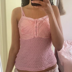 Rosa Topp - Supersöt rosa linnetopp med smockad nederdel och tunna axelband. Toppen har dekorativa knappar framtill och spetsdetaljer längs kanten. Perfekt för varma dagar och enkel att styla med jeans eller kjol.💕